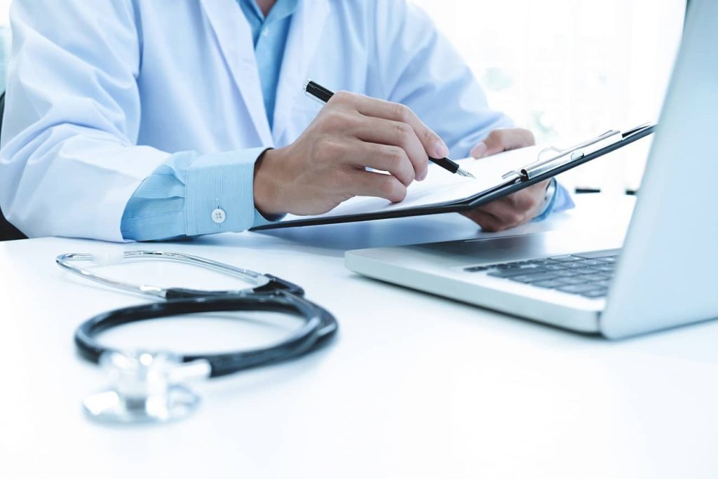 doctor-working-with-laptop-computer-writing-paperwork-hospital-background